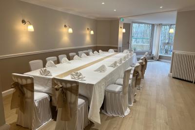 Salle lumineuse avec table ronde dressée pour un repas, nappes blanches et chaises décorées de rubans beiges.