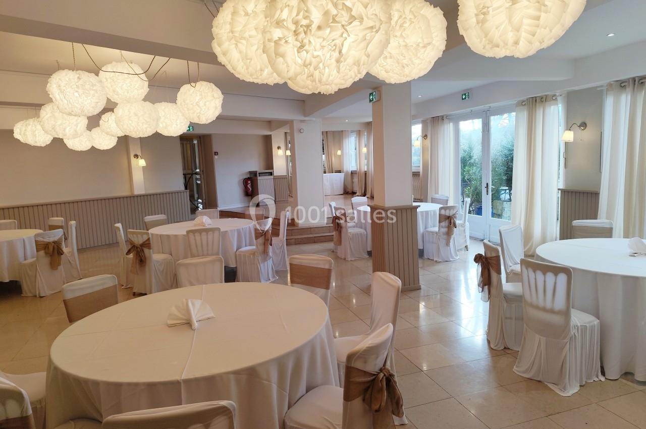Salle de réception lumineuse avec tables rondes dressées de nappes blanches et décorations suspendues en papier.