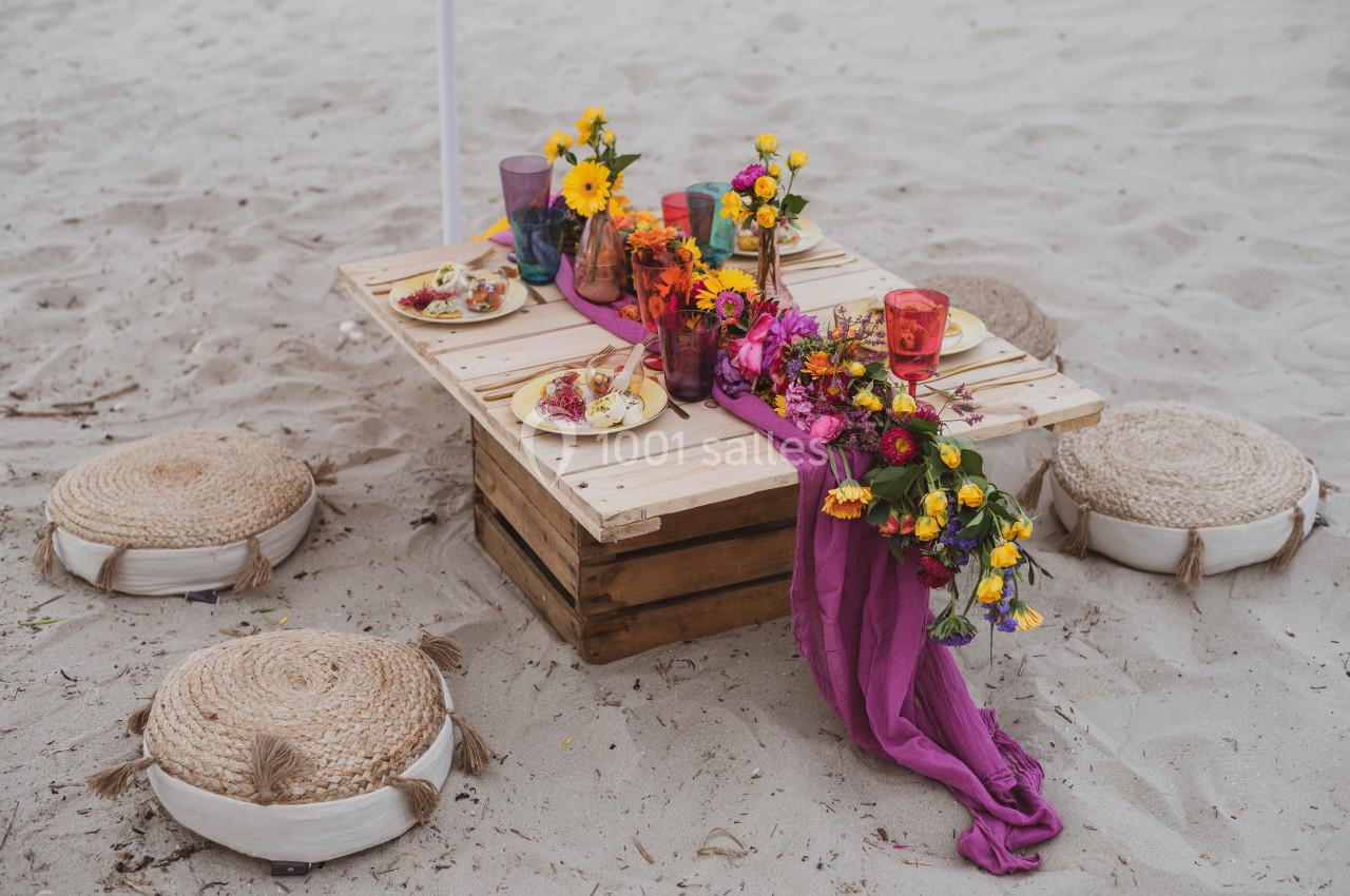 Table basse en bois décorée de fleurs colorées, serviettes violettes et vaisselle, entourée de coussins sur le sable.