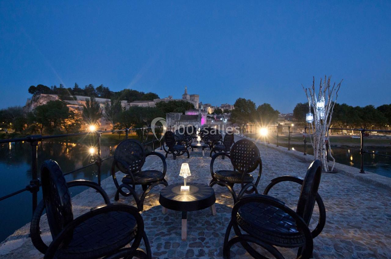 Terrasse extérieure avec tables et chaises noires, éclairée par des lampes, surplombant une rivière au crépuscule.