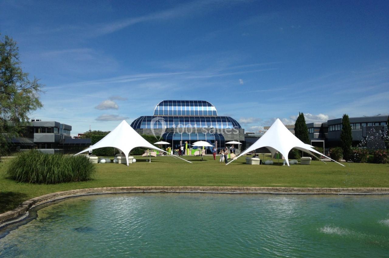 Pelouse avec tentes blanches modernes, bassin au premier plan et bâtiment vitré en arrière-plan sous un ciel bleu.