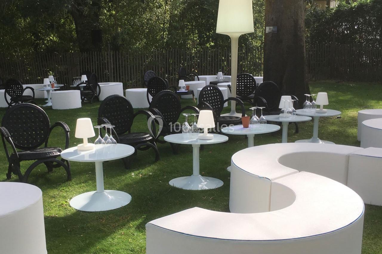 Mobilier de jardin avec tables blanches, chaises noires et lampes, disposé sur une pelouse entourée d'arbres.