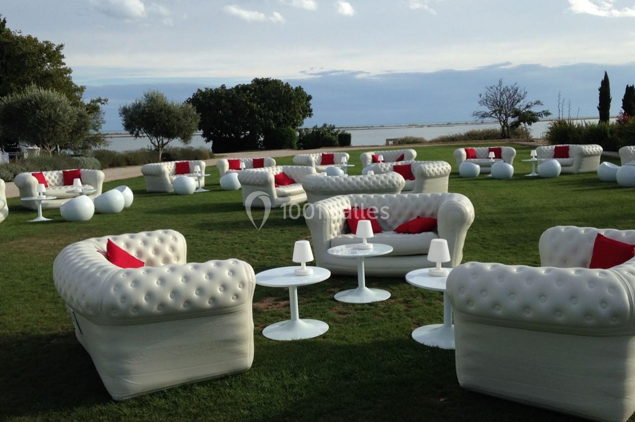 Mobilier de salon blanc avec coussins rouges disposé sur une pelouse en plein air, près d'un plan d'eau.