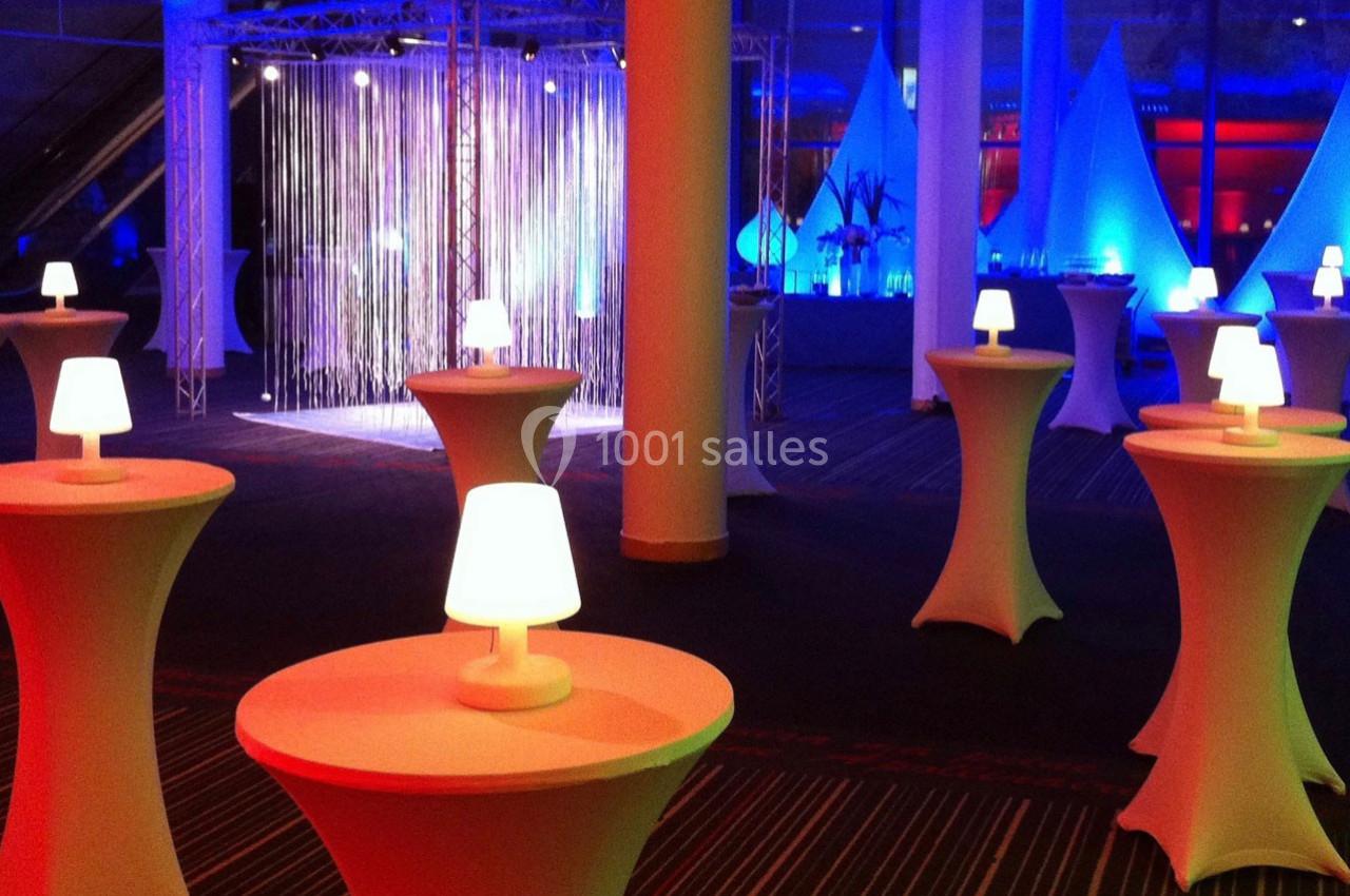 Salle décorée pour un événement avec des tables hautes éclairées et des lumières colorées en arrière-plan.