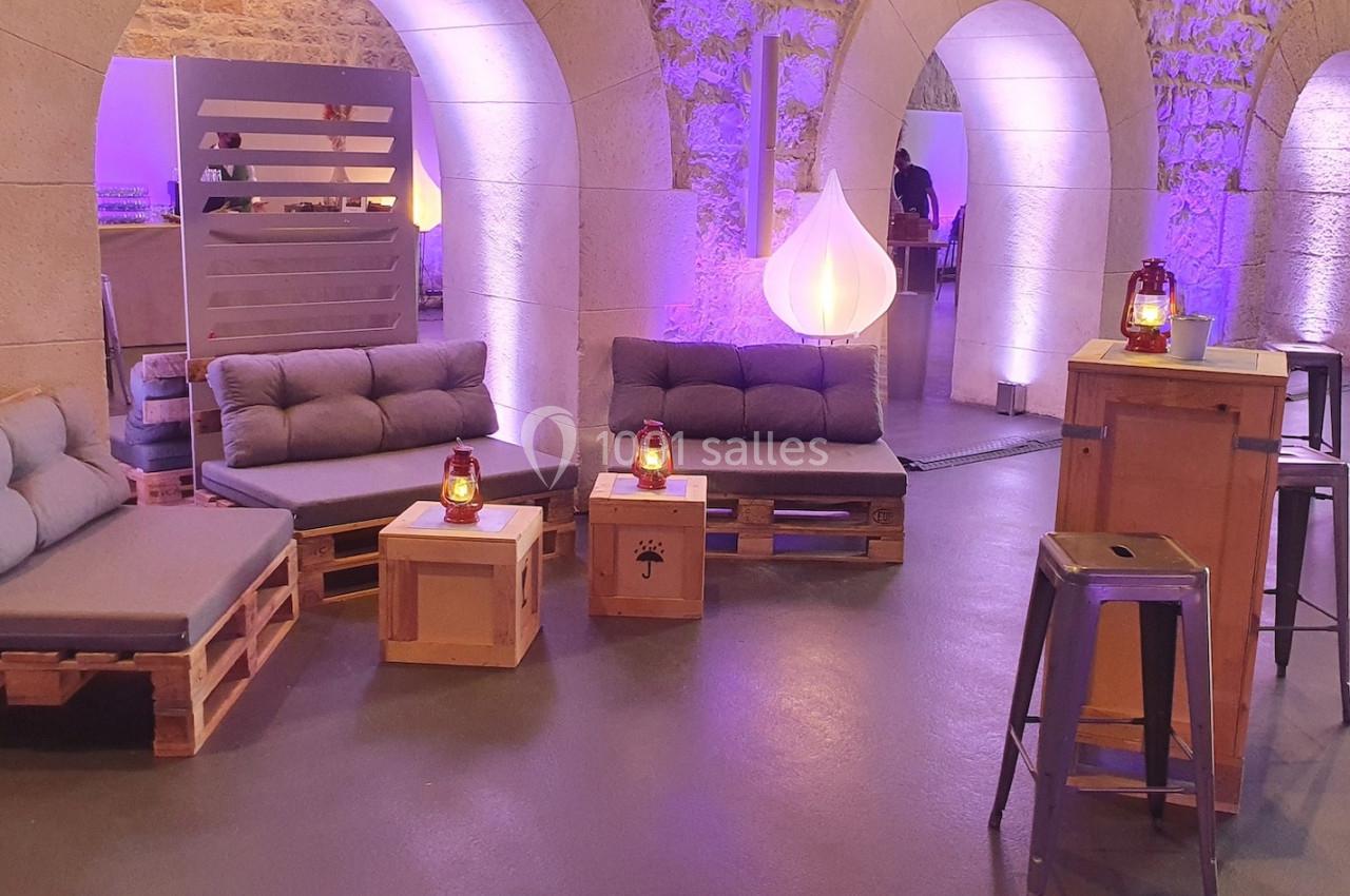 Espace lounge avec canapés en palettes, tables basses en bois et éclairage tamisé dans une salle voûtée en pierre.