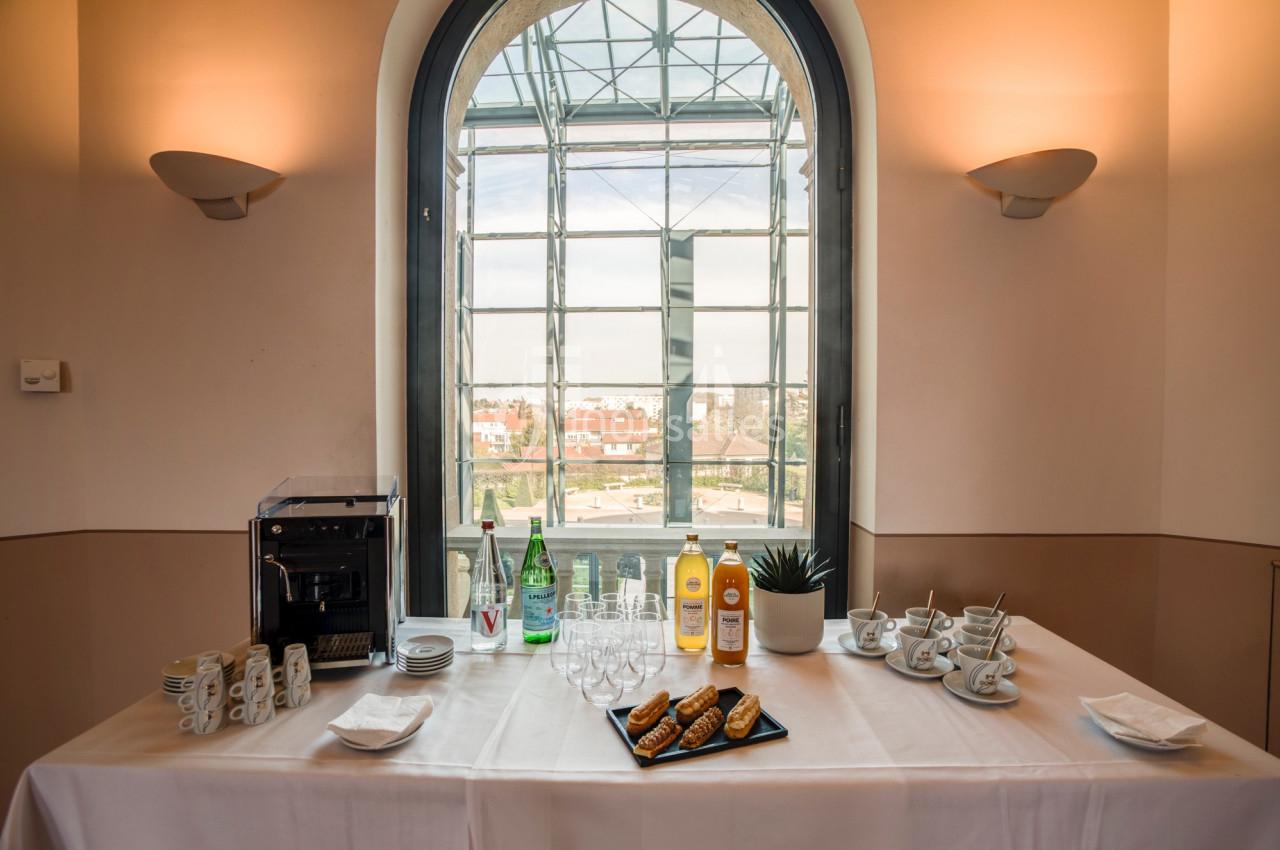 Location salle Saint-Priest (Rhône) - Château de Saint Priest #14 Table dressée avec machine à café, bouteilles, verres, tasses et viennoiseries, devant une grande fenêtre lumineuse.