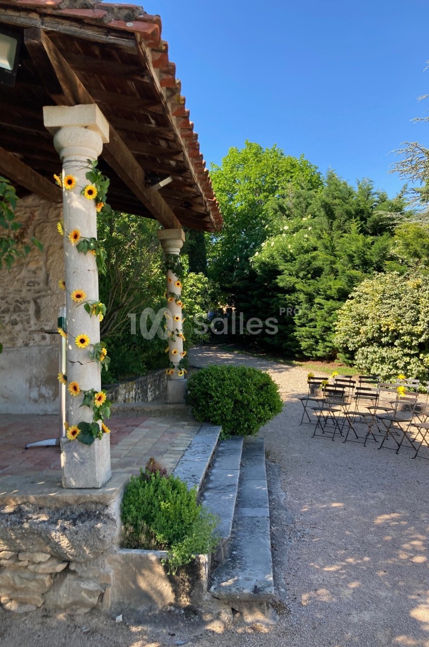 Terrasse en pierre avec colonnes décorées de tournesols, entourée de verdure, et chaises disposées en extérieur.