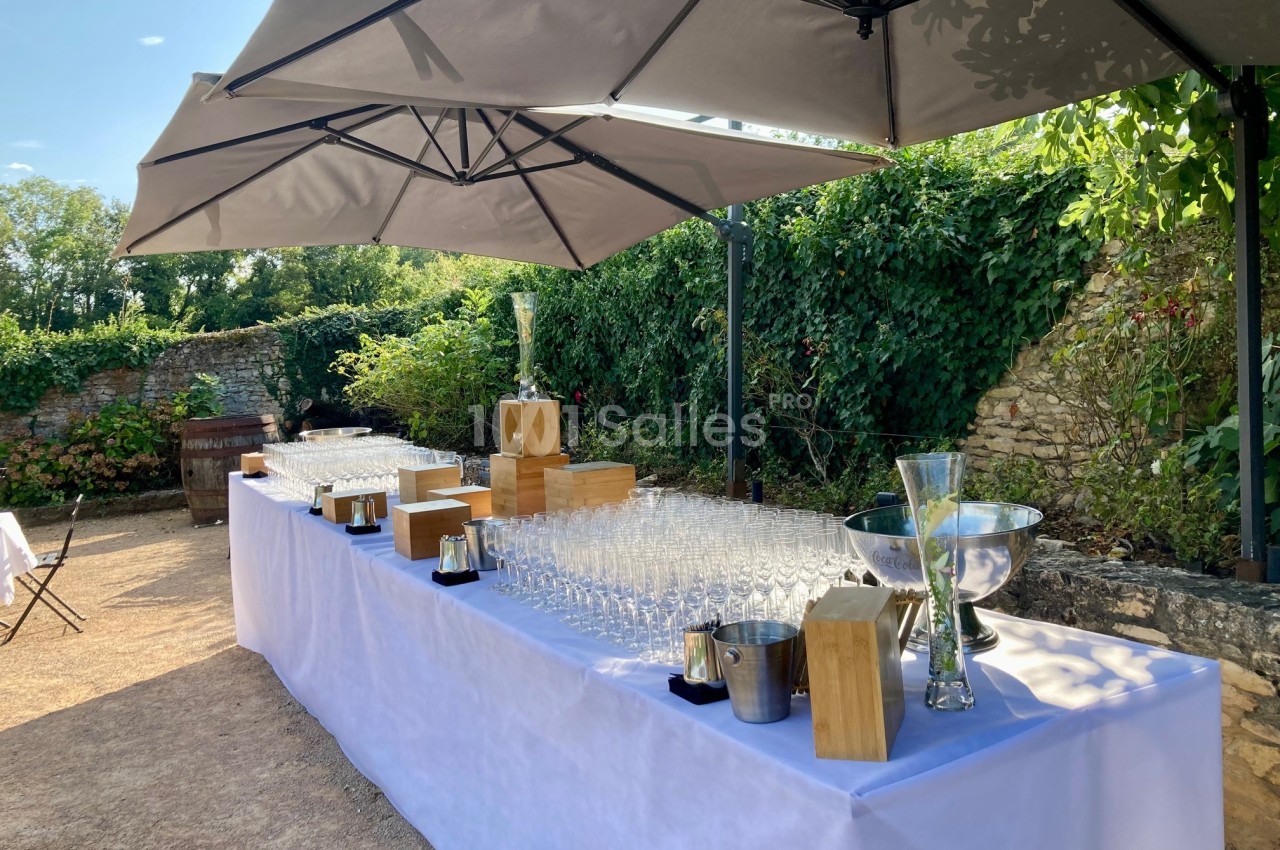 Table dressée en extérieur avec des verres, seaux à glace et décorations sous des parasols, entourée de verdure.
