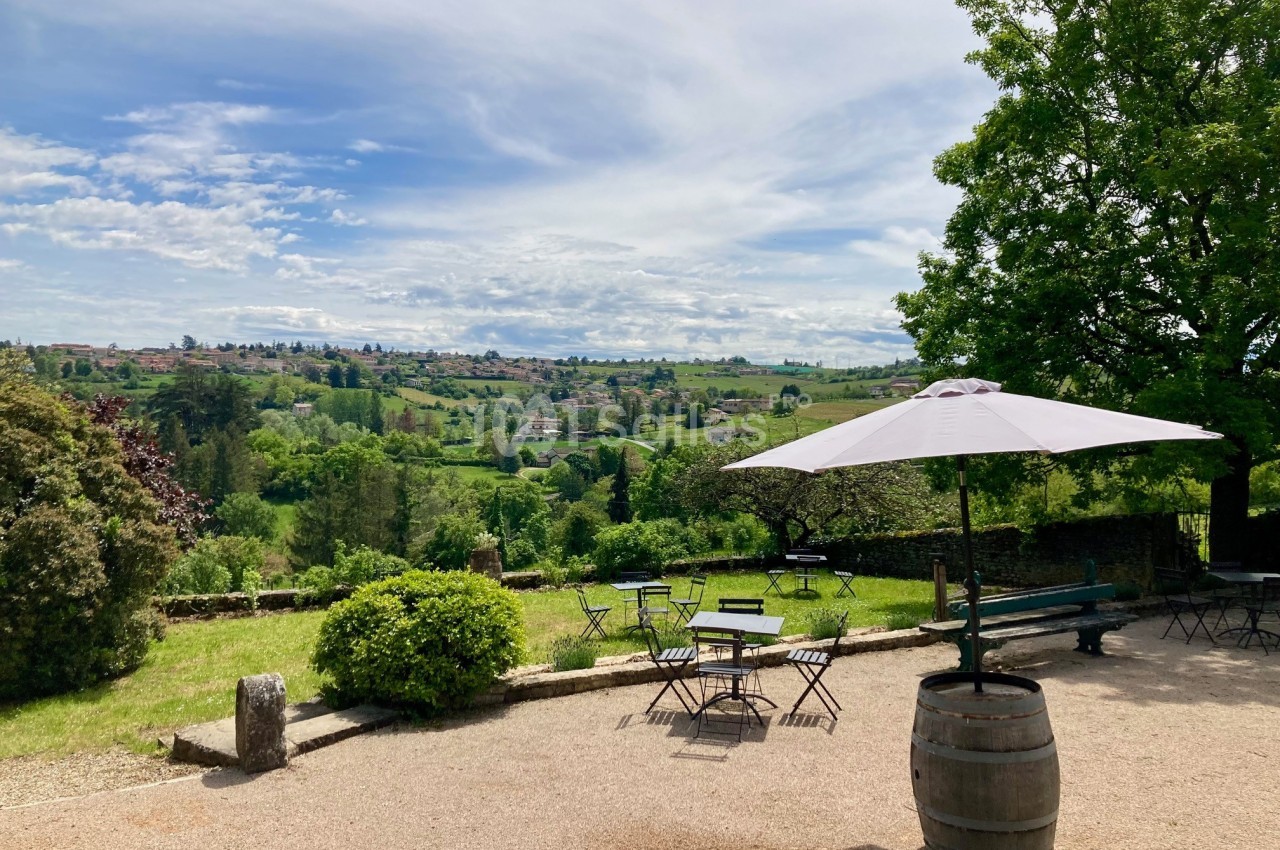 Terrasse avec tables et parasol, offrant une vue dégagée sur un paysage vallonné et verdoyant sous un ciel partiellement…