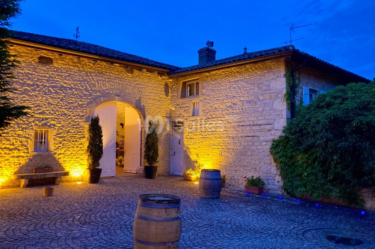 Cour pavée éclairée au crépuscule, entourée de murs en pierre, avec des tonneaux et des plantes décoratives.