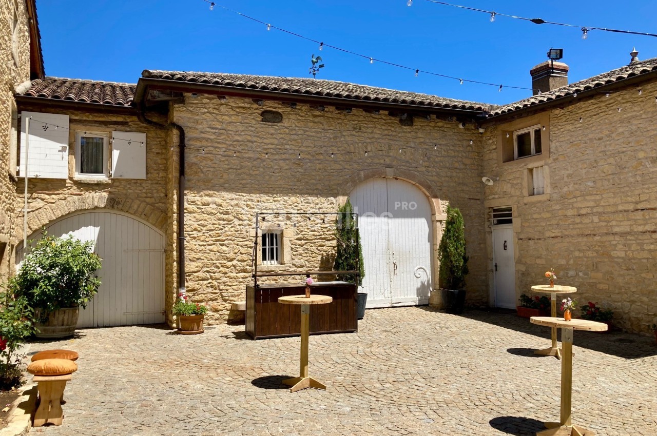 Cour pavée entourée de murs en pierre, avec des tables hautes, des plantes et des guirlandes lumineuses suspendues.