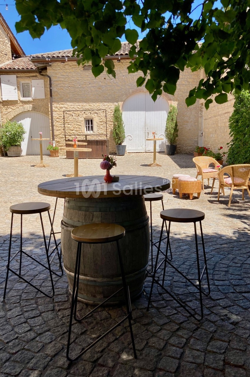 Table haute en tonneau entourée de tabourets sur une cour pavée, avec des bâtiments en pierre en arrière-plan.