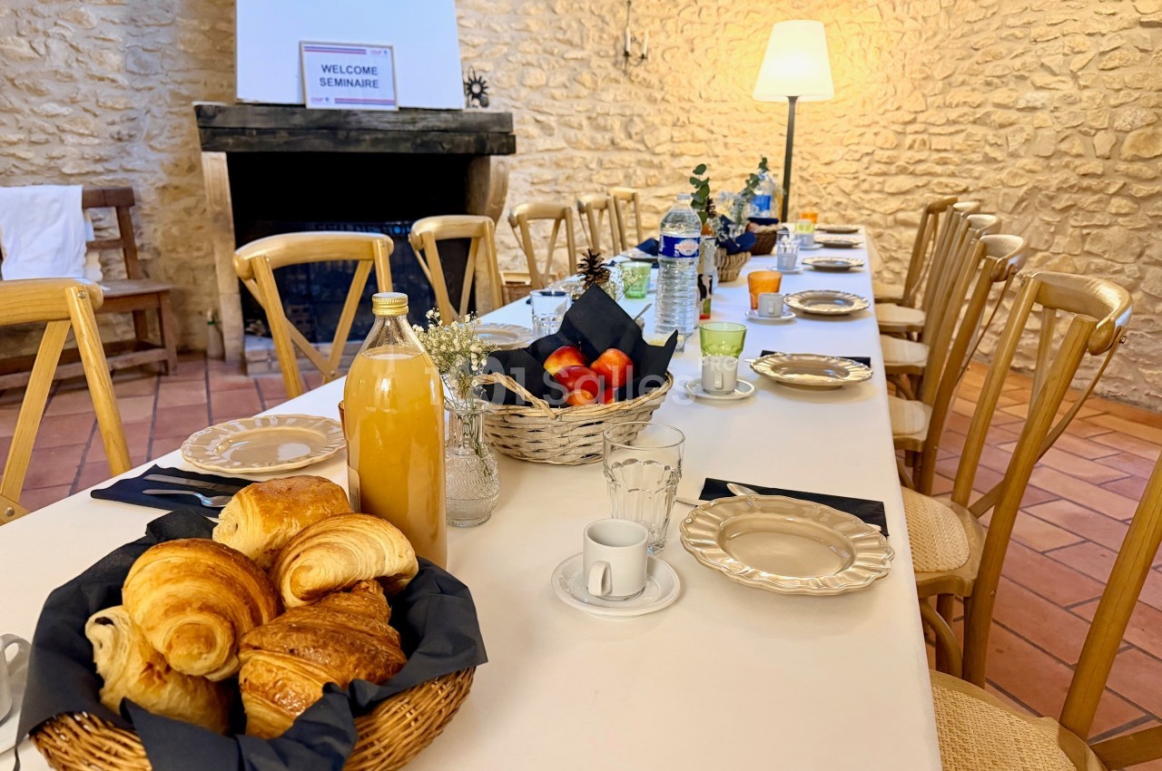 Table dressée pour un petit-déjeuner avec viennoiseries, fruits, jus et café, dans une salle en pierre chaleureuse.