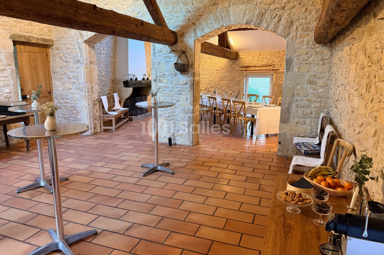Salle en pierre avec poutres apparentes, tables dressées, chaises en bois et buffet avec fruits et boissons.
