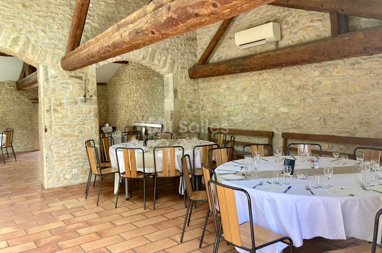 Salle en pierre avec poutres apparentes, tables rondes dressées avec nappes blanches et chaises en bois.
