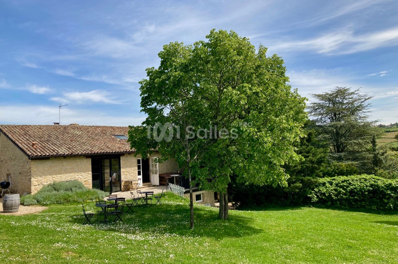 Maison en pierre avec toit en tuiles, entourée d'un jardin verdoyant, un arbre au premier plan et ciel dégagé.