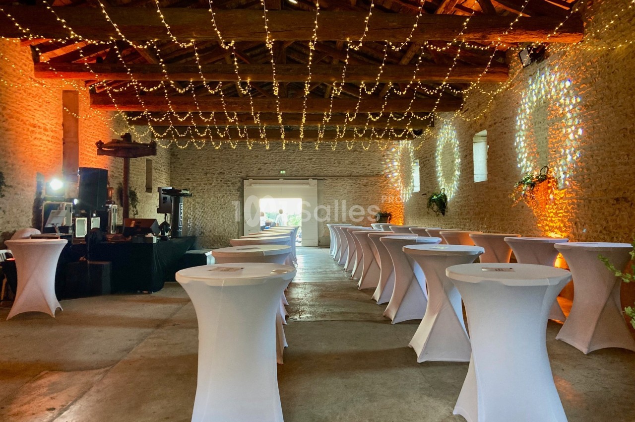 Grande salle en pierre avec tables hautes recouvertes de nappes blanches, éclairée par des guirlandes lumineuses suspendues.