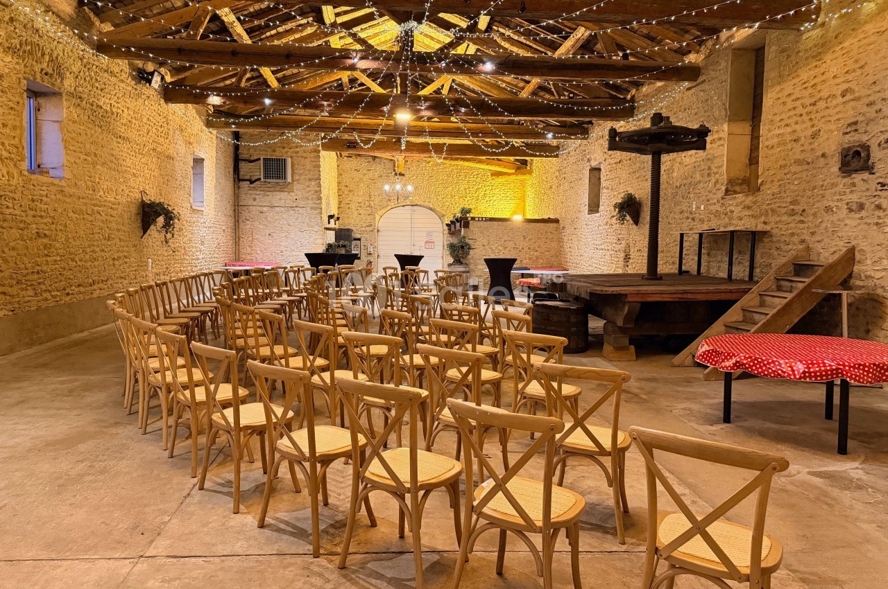 Salle en pierre avec des chaises en bois disposées face à une petite scène, éclairée par des guirlandes lumineuses.