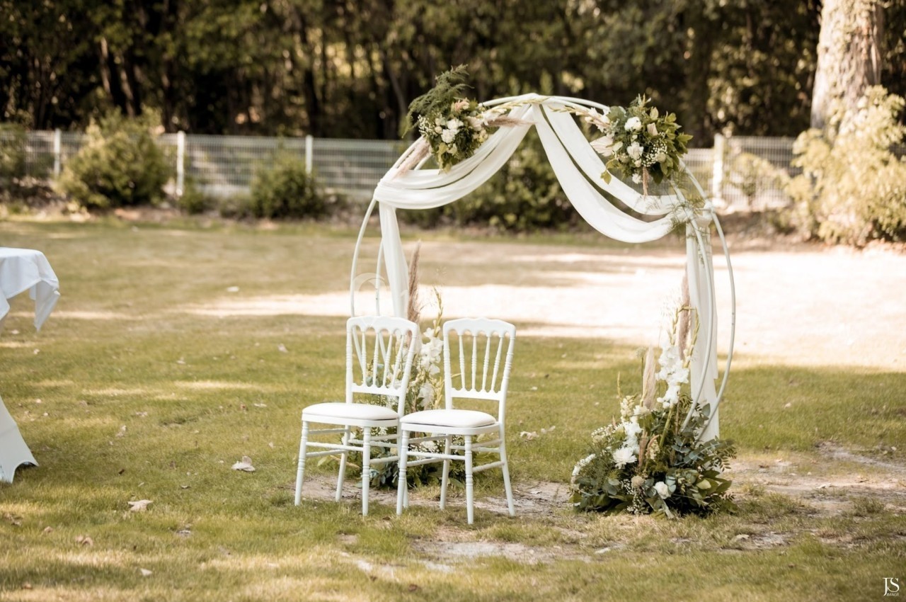 Arche de mariage décorée de fleurs blanches et de voilages, avec deux chaises blanches sur une pelouse.