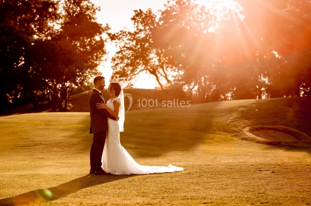Un couple en tenue de mariage se tient enlacé sur une pelouse au coucher du soleil, entouré d'arbres.