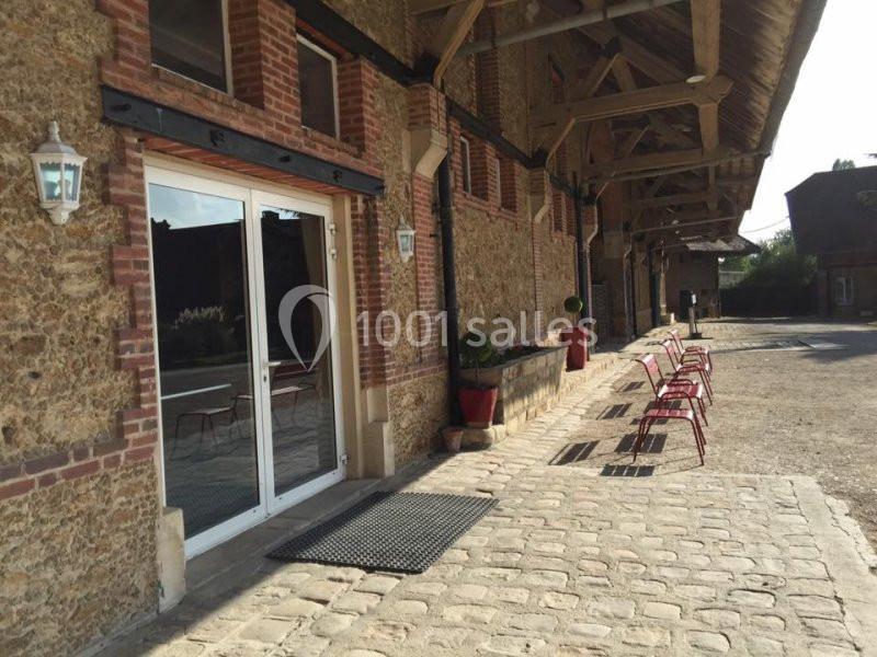 Façade en pierre et briques avec une entrée vitrée, des chaises rouges alignées et une cour pavée ensoleillée.