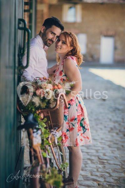 Un couple souriant près d'un vélo décoré de fleurs, dans une ruelle pavée et ensoleillée.