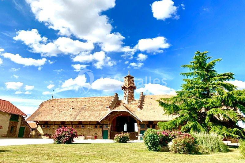 Bâtiment ancien en pierre avec un clocher, entouré de pelouse, d'arbustes fleuris et d'un ciel bleu parsemé de nuages.