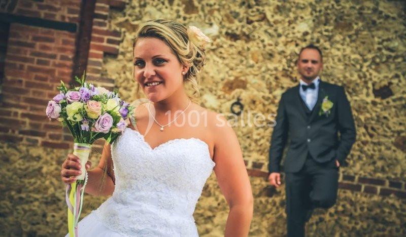 Une mariée en robe blanche tient un bouquet de fleurs devant un mur en pierre, un homme en costume en arrière-plan.