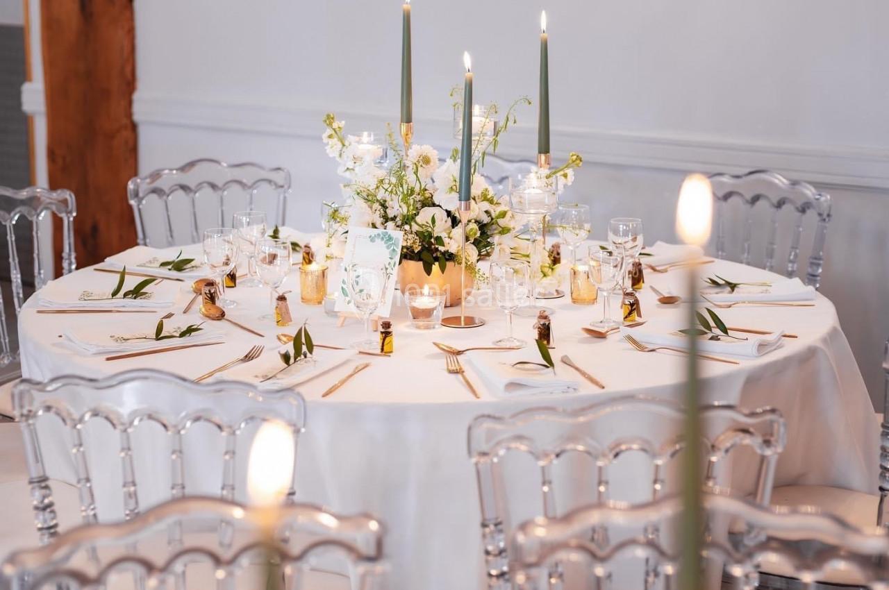 Table ronde décorée pour un événement, avec nappes blanches, bougies allumées et chaises transparentes.