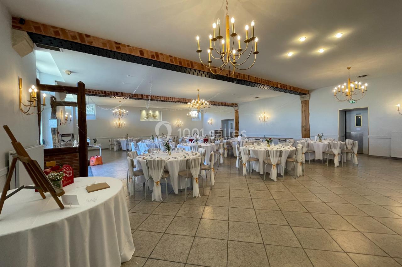 Salle de réception lumineuse avec tables rondes dressées, décorée de lustres et poutres apparentes.