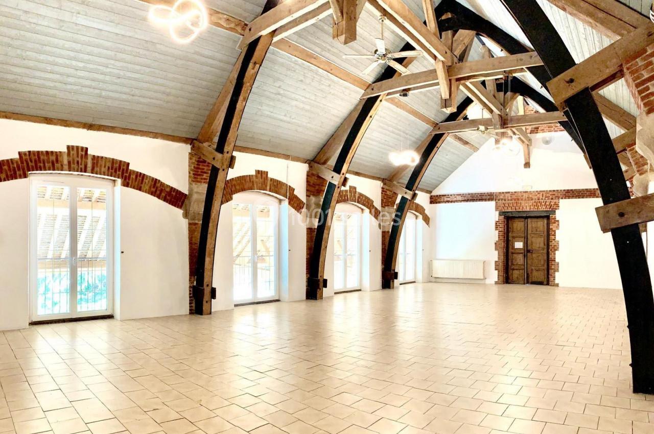 Salle spacieuse avec poutres apparentes, murs en briques et grandes fenêtres laissant entrer la lumière naturelle.