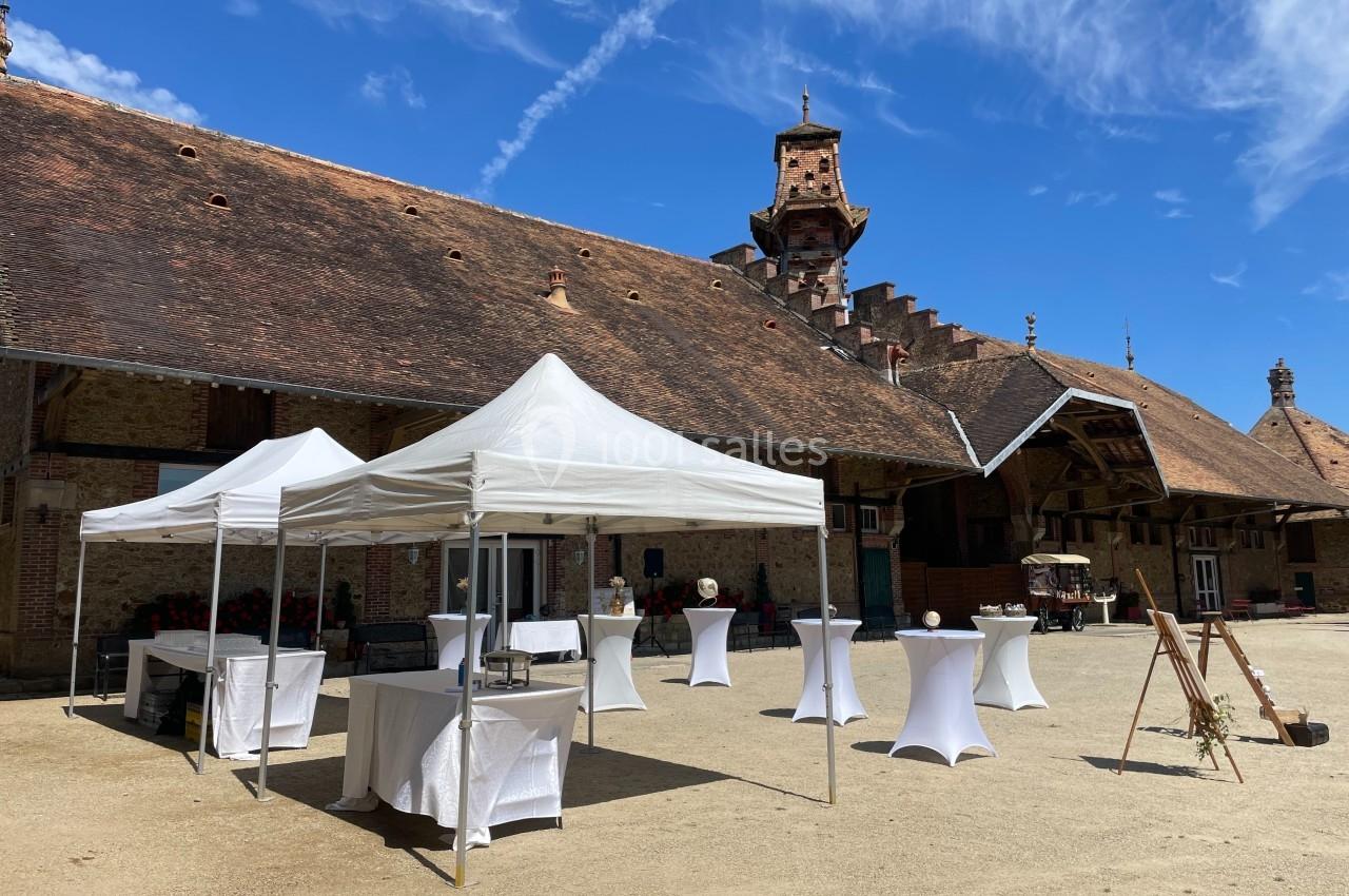 Cour extérieure avec des tentes blanches, tables hautes et bâtiment ancien en arrière-plan sous un ciel bleu.