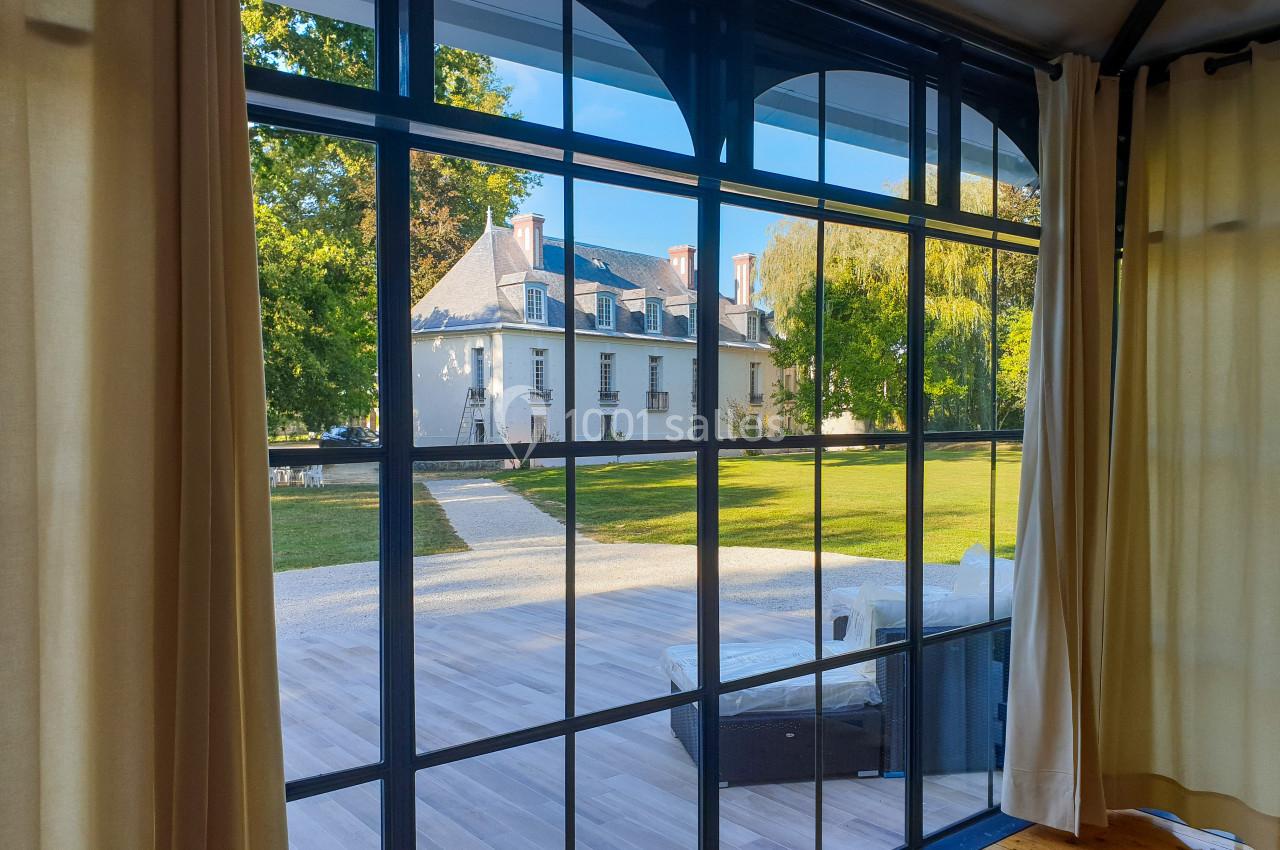 Location salle Fontains (Seine-et-Marne) - Château de Bourguignon #6 Vue sur un jardin et une maison ancienne à travers une grande baie vitrée avec des rideaux beiges ouverts.