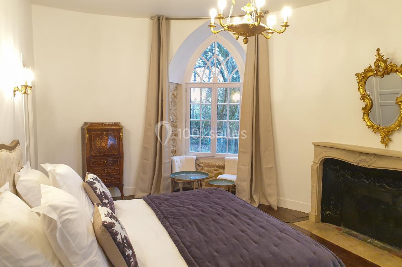 Location salle Fontains (Seine-et-Marne) - Château de Bourguignon #25 Chambre lumineuse avec lit double, cheminée en pierre, miroir doré, meuble ancien et grande fenêtre en arche.