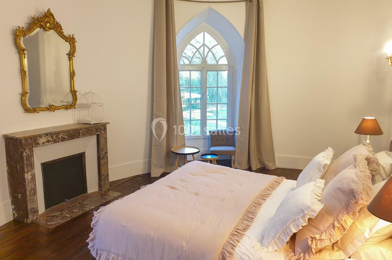 Location salle Fontains (Seine-et-Marne) - Château de Bourguignon #26 Chambre lumineuse avec lit double, cheminée en marbre, miroir doré et grande fenêtre en arc avec rideaux beiges.