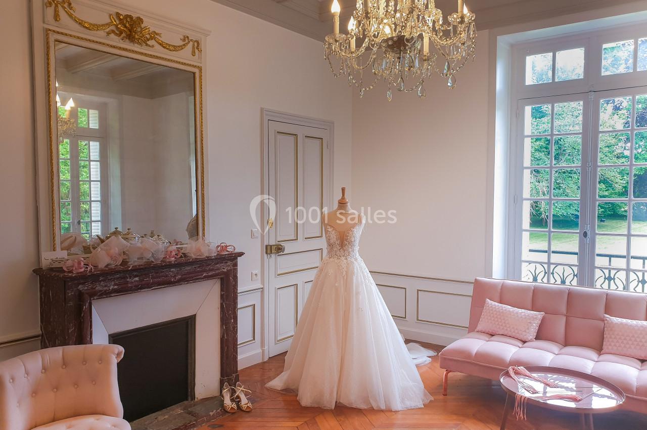 Location salle Fontains (Seine-et-Marne) - Château de Bourguignon #5 Robe de mariée exposée sur un mannequin dans un salon élégant avec cheminée, lustre et grandes fenêtres donnant sur un…