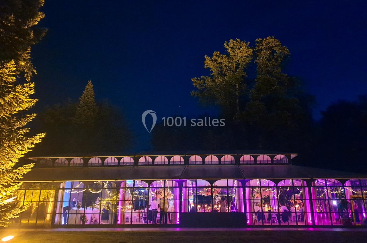 Location salle Fontains (Seine-et-Marne) - Château de Bourguignon #33 Vue nocturne d'une verrière illuminée par des lumières violettes, entourée d'arbres et d'un jardin.