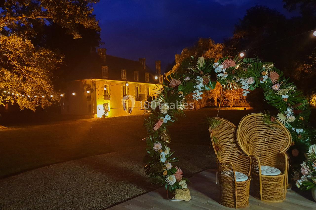 Location salle Fontains (Seine-et-Marne) - Château de Bourguignon #9 Arc floral illuminé avec deux fauteuils en rotin devant un bâtiment historique éclairé, de nuit.