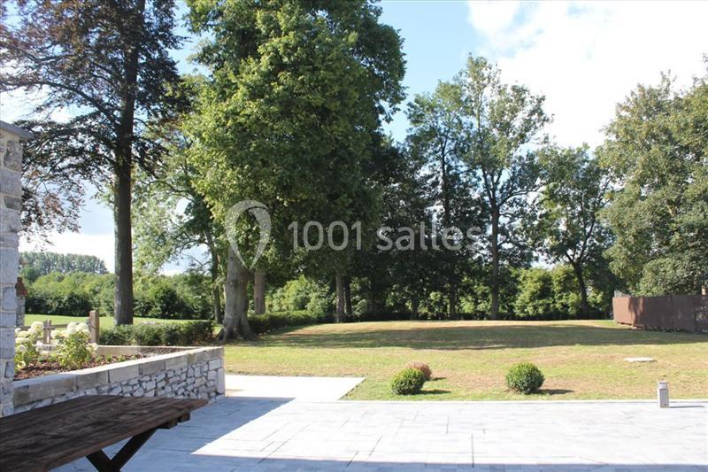 Vue d'un jardin avec pelouse, arbres et terrasse en pierre, sous un ciel partiellement dégagé.