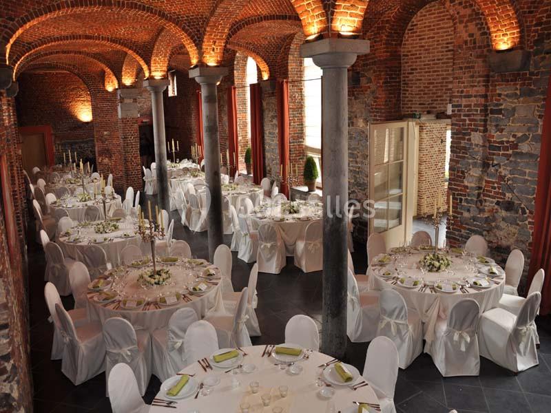 Salle voûtée en briques avec des tables rondes décorées pour un événement, chaises blanches et lumière tamisée.