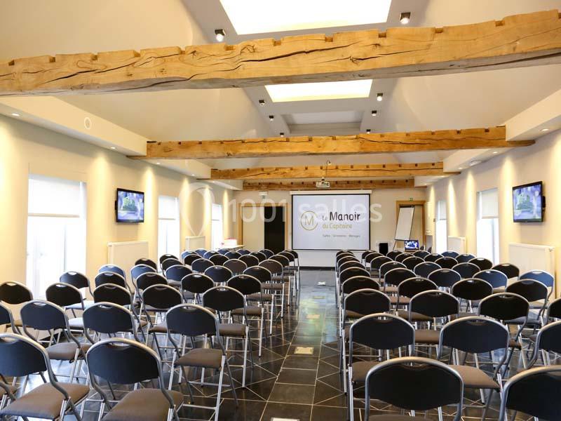 Salle de conférence lumineuse avec poutres apparentes, rangées de chaises noires et écran de projection à l'avant.