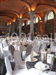 Salle voûtée en briques avec tables rondes dressées pour un événement, nappes blanches et serviettes pliées.