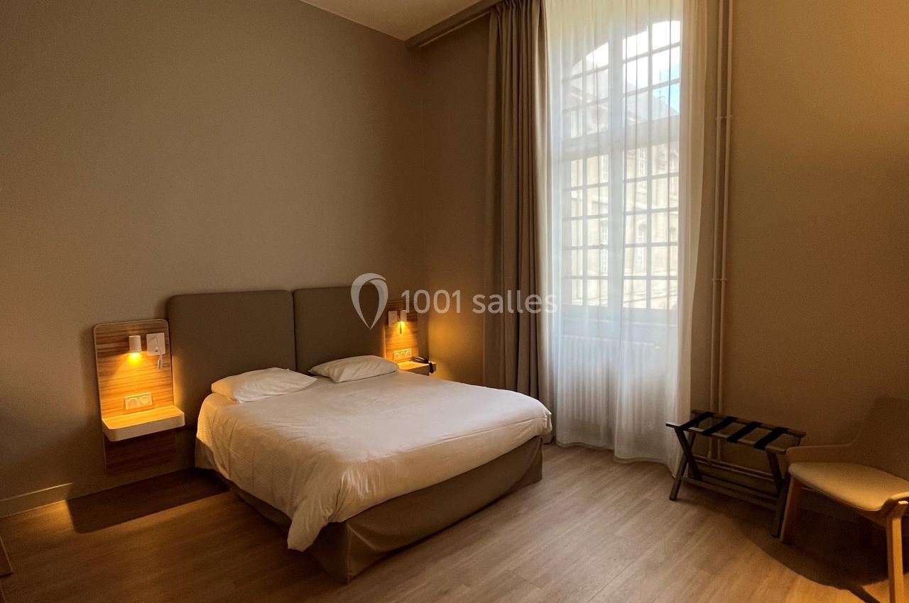 Chambre avec lit double, chevets éclairés, fauteuil, porte-bagages et grande fenêtre avec rideaux translucides.