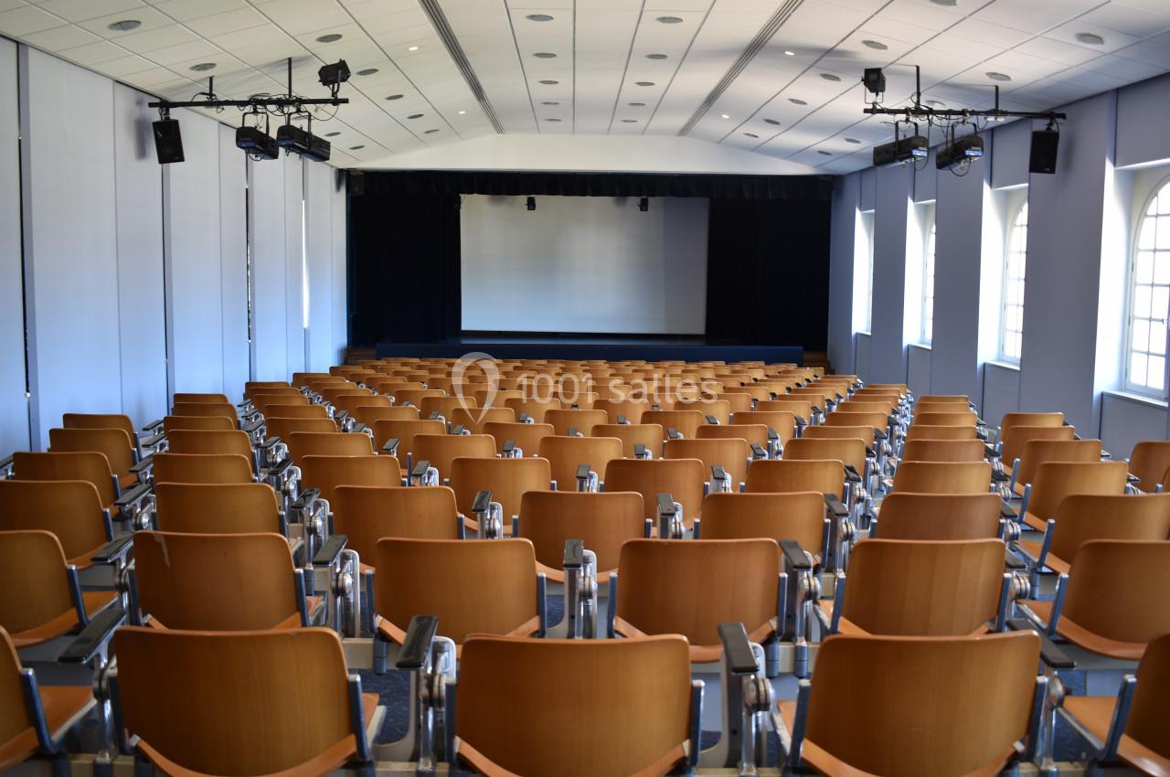 Salle de conférence vide avec des rangées de sièges en bois, un écran blanc et des projecteurs au plafond.
