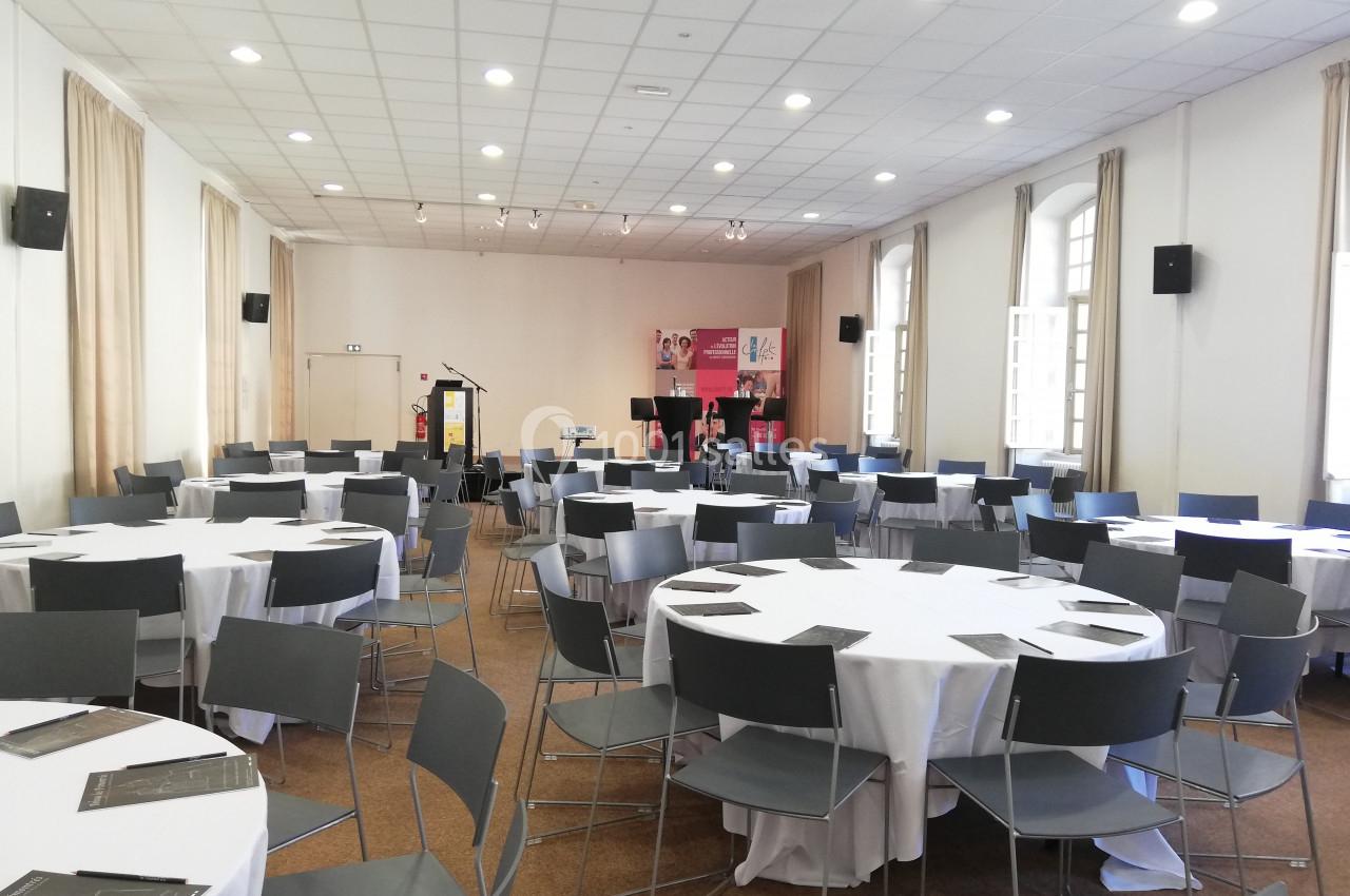 Salle de conférence lumineuse avec des tables rondes dressées de nappes blanches et des chaises grises.
