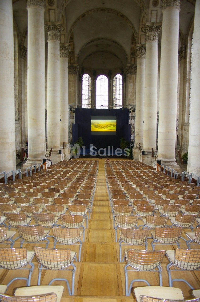 Salle avec de hautes colonnes, rangées de chaises en métal et un écran de projection au fond.