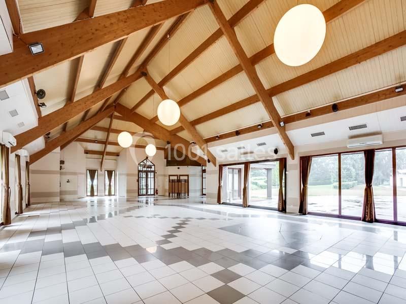 Grande salle lumineuse avec charpente en bois apparente, carrelage clair et larges baies vitrées donnant sur l'extérieur.