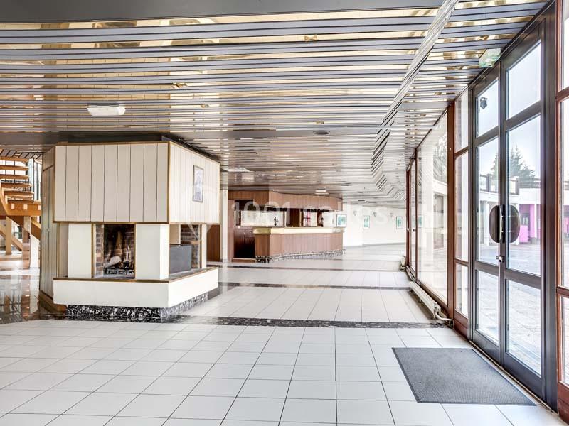 Intérieur lumineux avec sol carrelé, cheminée moderne, comptoir en bois et grandes baies vitrées donnant sur l'extérieur.