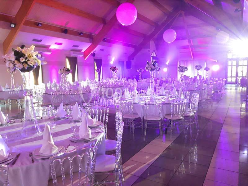 Salle de réception décorée avec des tables dressées, nappes blanches, chaises transparentes et éclairage violet.