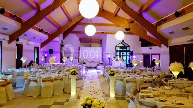 Salle de réception décorée pour un mariage, avec tables dressées, éclairage violet et allée centrale ornée de pétales.